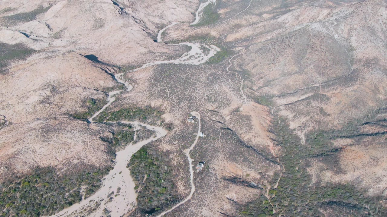 Drone frame over Cabo Pulmo's dry ridges where a pale road curves through scrub and rocky ground, with no visible people.
