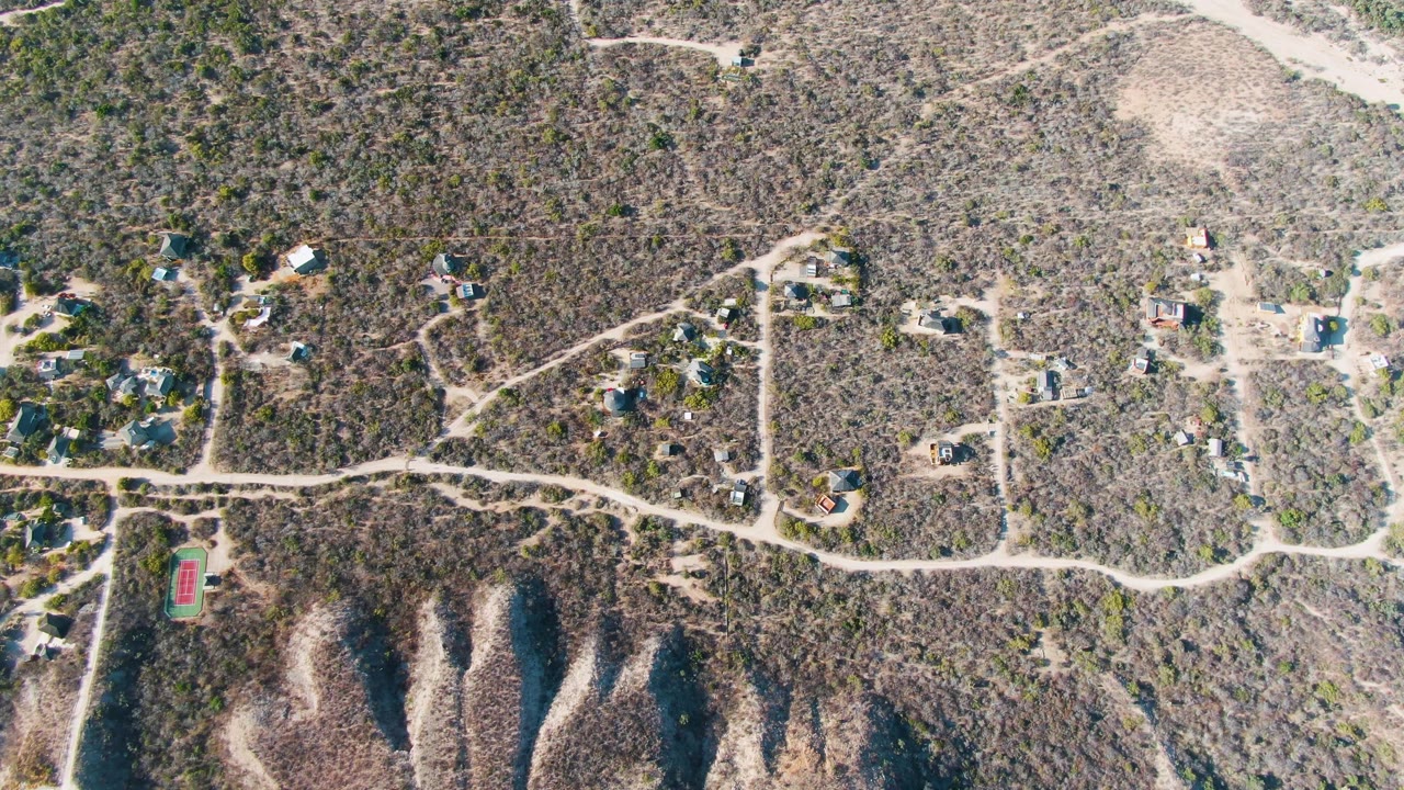Static aerial frame over scattered hillside homes and pale dirt roads in Cabo Pulmo, Baja California Sur, with dry vegetation and no visible people.