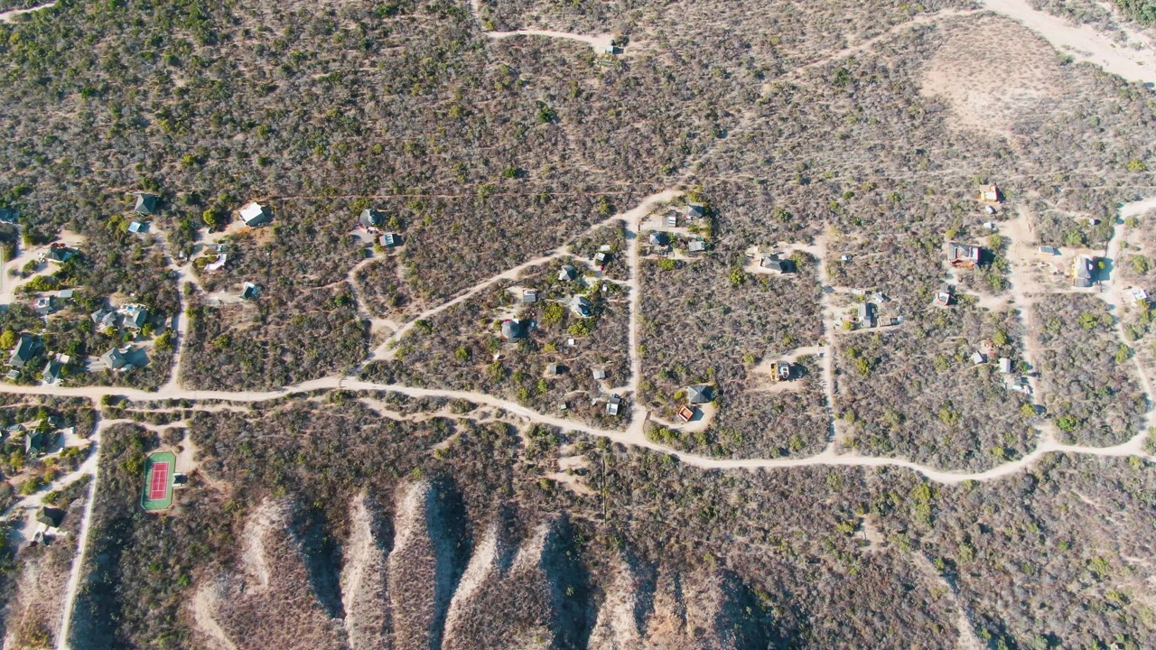 High aerial view over Cabo Pulmo hillside homes, pale dirt roads, dry scrub vegetation, and a red rectangular court-like structure near the lower left, with no visible people.