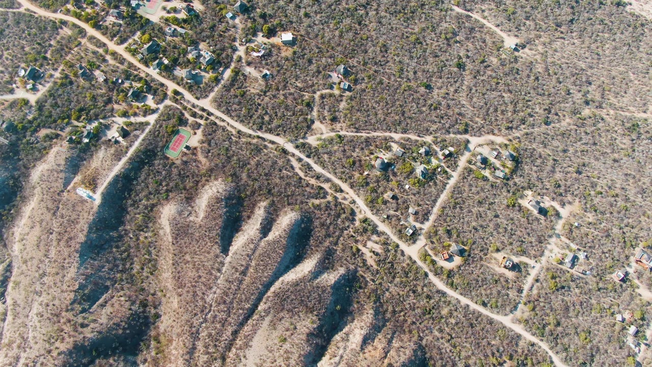 High aerial view over Cabo Pulmo, Baja California Sur, showing winding dirt roads, sparse hillside vegetation, scattered homes, dry gullies, and no visible people at the start of the clip.