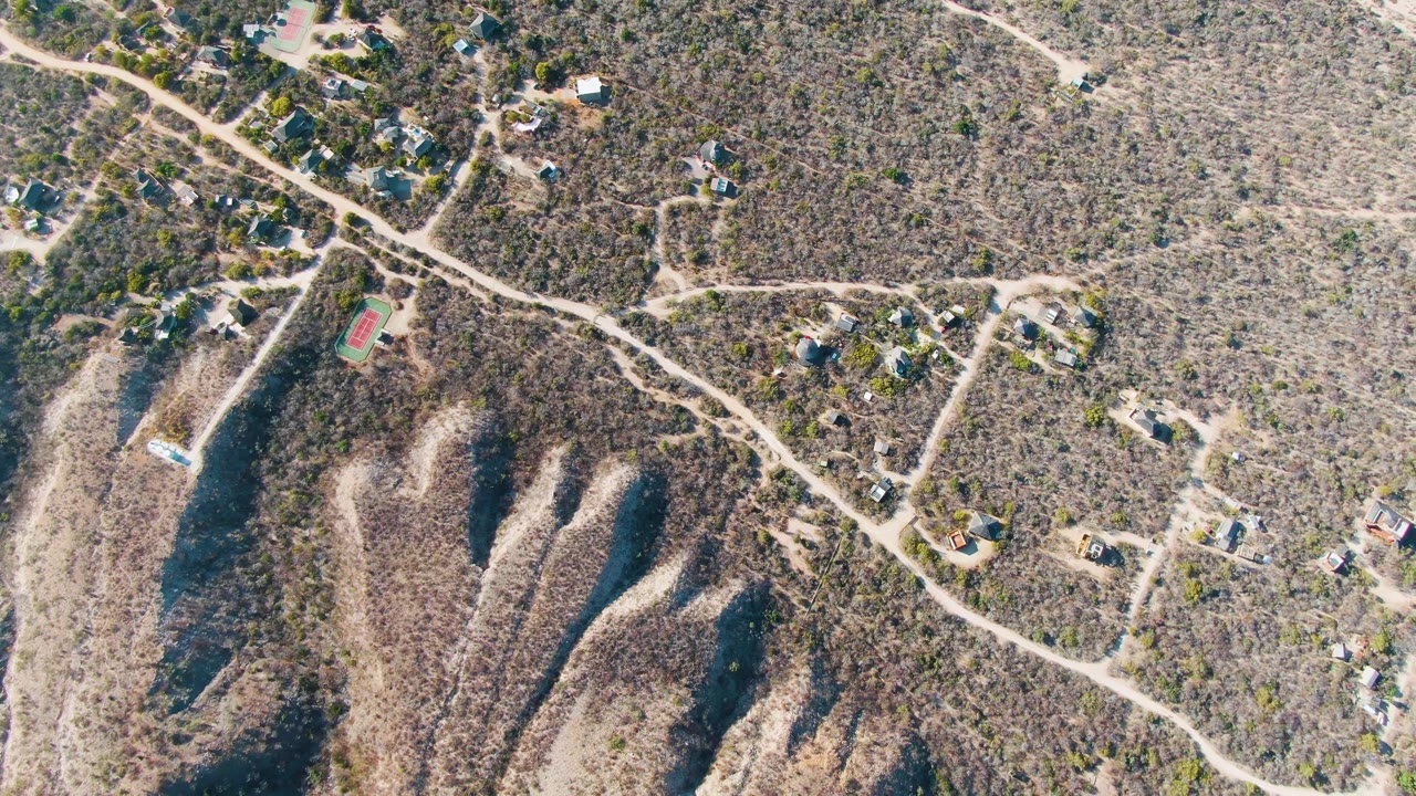 Aerial frame over Cabo Pulmo, Baja California Sur, with pale dirt tracks crossing scrubland, homes grouped near the upper edge, shadowed ravines below, and no visible people.