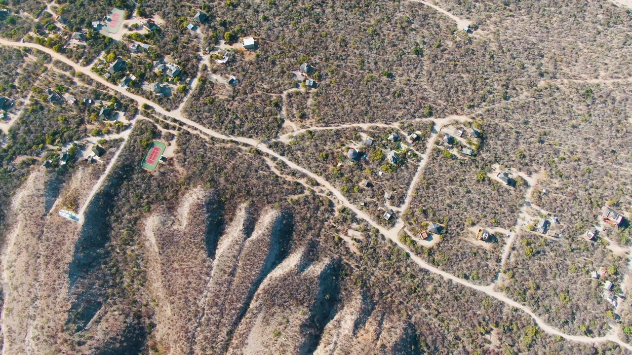 Drone-style overhead view in Cabo Pulmo, Baja California Sur, showing clustered houses, an unpaved road curving across dry brush, a small colored court, and no visible people.