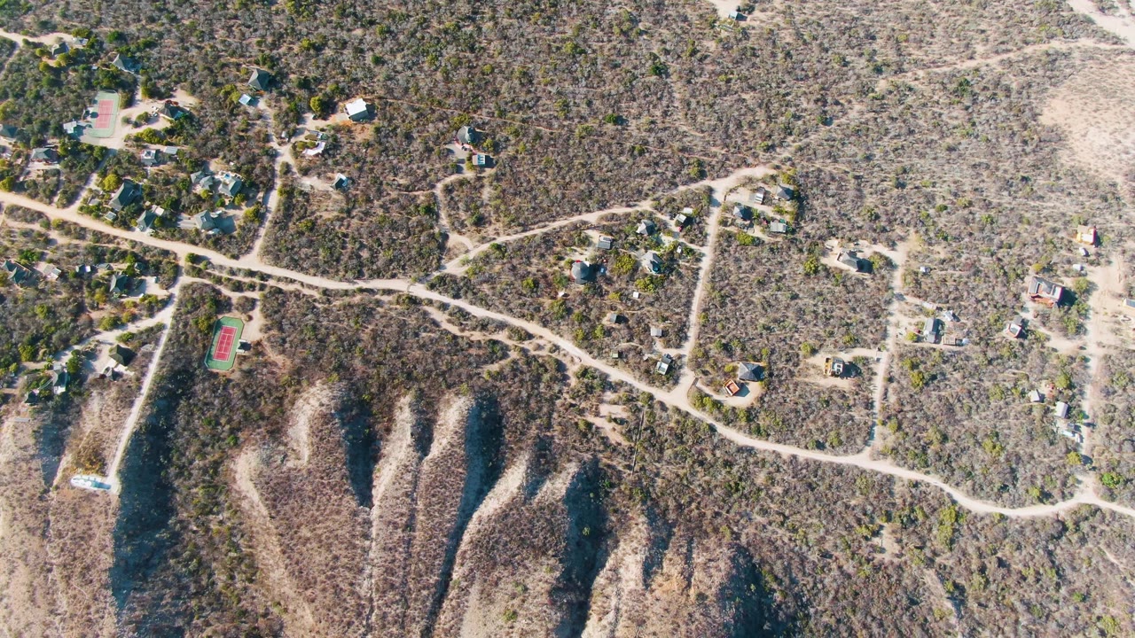 High aerial frame over Cabo Pulmo, Baja California Sur showing scattered homes, pale winding roads, dry scrub, ridge shadows, and no visible people.