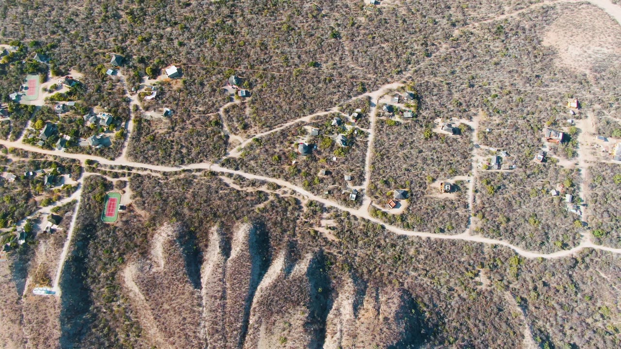 Drone frame above Cabo Pulmo showing homes set back from pale dirt roads, rough desert vegetation, eroded ridge forms, and no visible people.