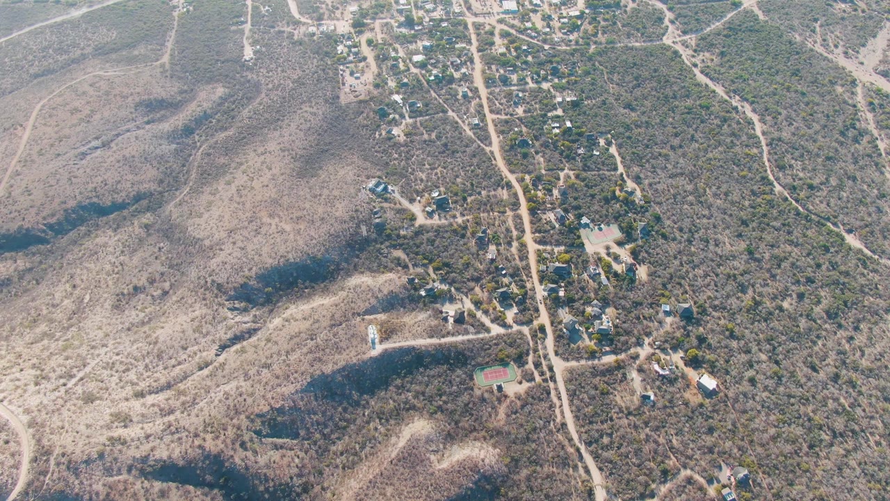 Aerial frame from Cabo Pulmo, Baja California Sur, showing a car on a pale dirt road between scrub-covered hills and scattered buildings, with no visible people.
