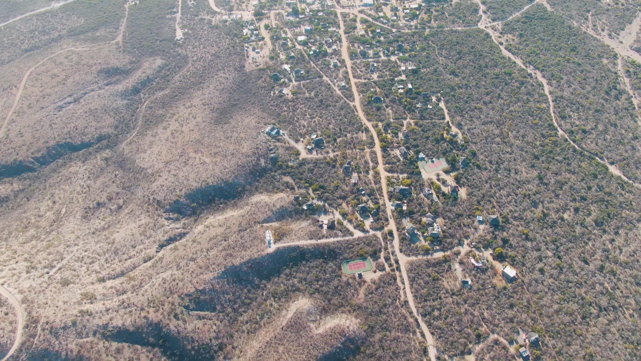 Drone view of a dirt road beside scrubby hills and scattered buildings.