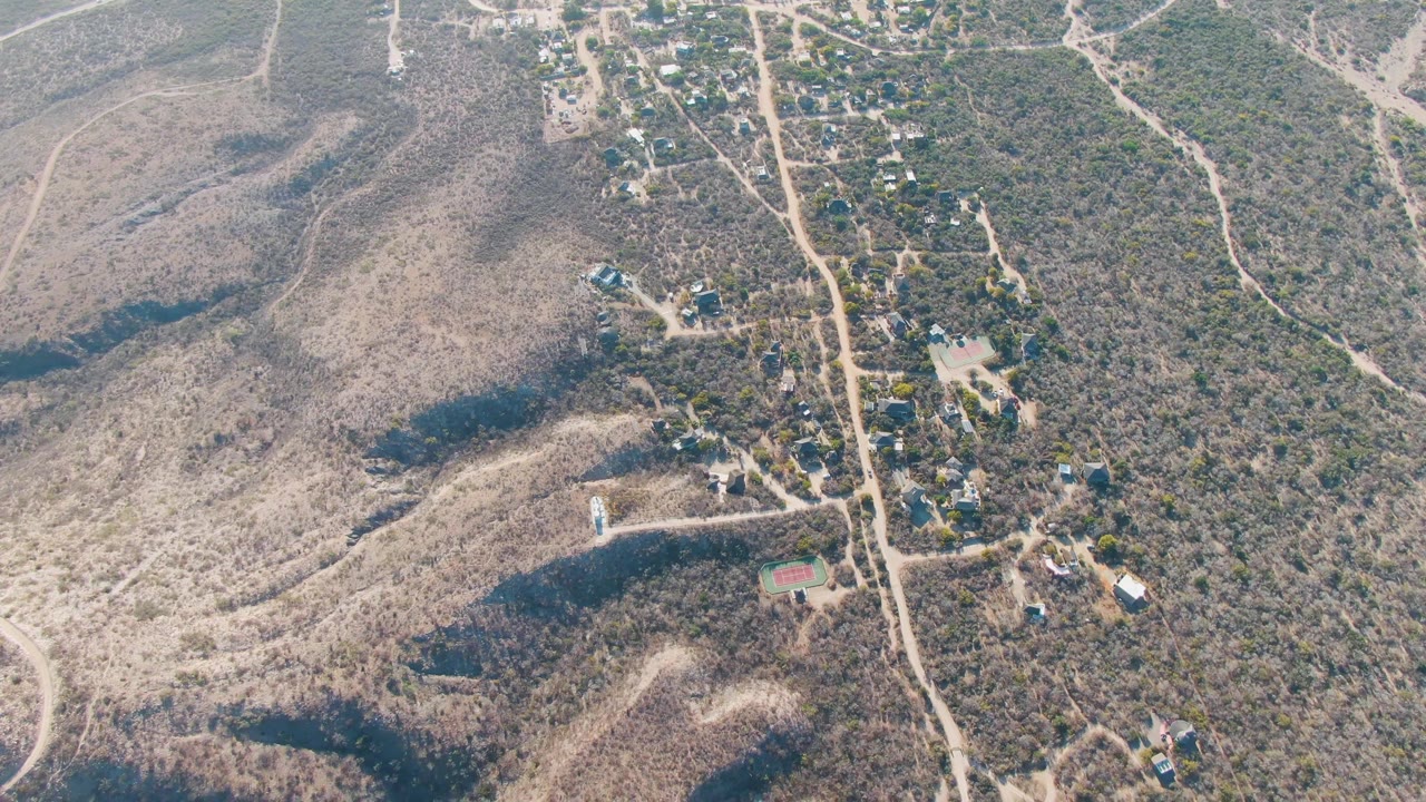 Aerial frame from Cabo Pulmo, Baja California Sur, showing the road, clustered hillside buildings, dry brush, pale slopes, and a small vehicle on the route with no visible people.