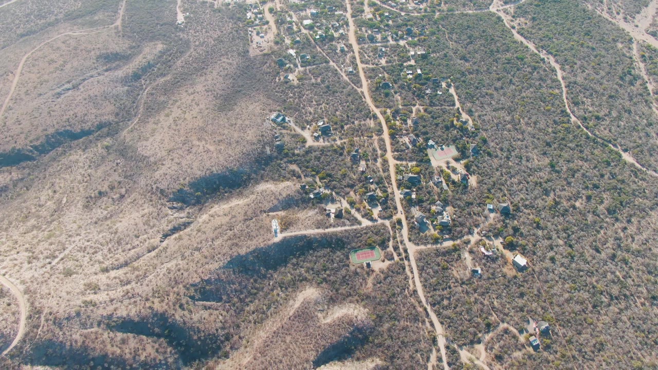 Aerial view of dry hills, a dirt road, and scattered homes with a car present.