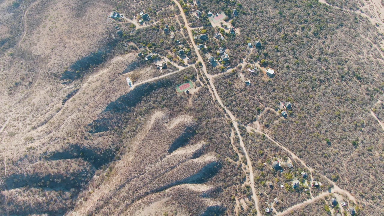 Drone-style overhead frame near Cabo Pulmo with winding dirt tracks, scattered hillside structures, dry brush, shaded gullies, and no visible people.
