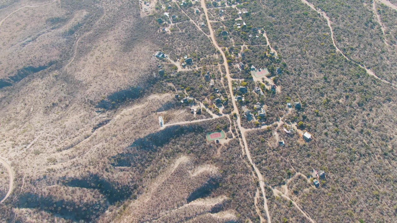 High aerial frame over dry hills and scattered houses near Cabo Pulmo, Baja California Sur, with pale dirt roads crossing scrub terrain and no visible people.