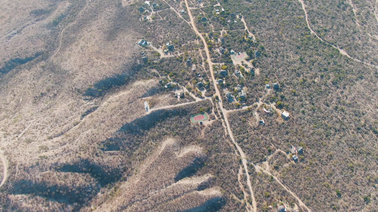 High drone frame over Cabo Pulmo area hillside terrain, with clustered homes near winding dirt tracks, tan scrub, and no visible people.