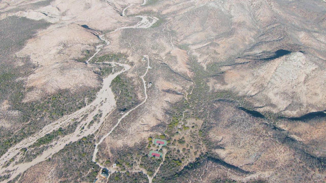 Aerial view over Cabo Pulmo dry hills with a pale winding dirt road, sparse scrub, a small red-roofed settlement near the lower edge, and no visible people.
