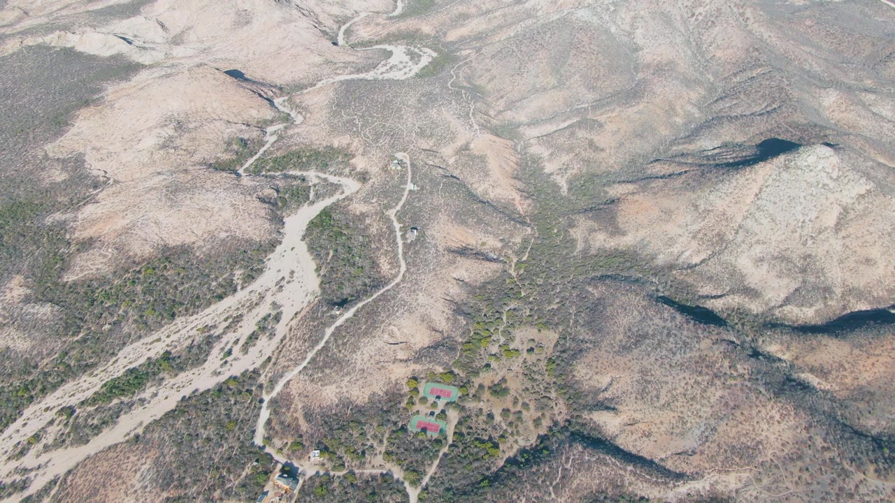 High aerial frame over Cabo Pulmo arid hills with a winding dirt track, pale drainage lines, scattered green vegetation, red-roofed structures low in frame, and no visible people.