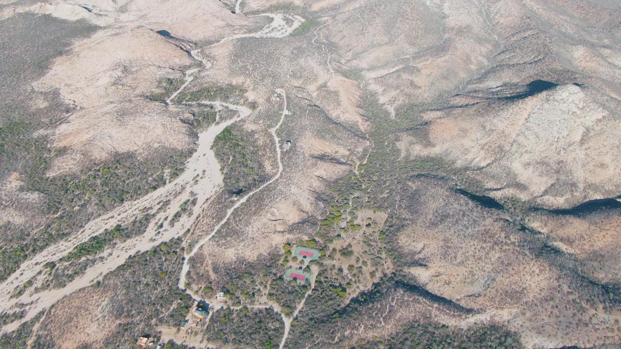 High aerial frame over Cabo Pulmo dry hills shows a winding dirt road, sparse vegetation, rocky ridges, a few small buildings near the lower frame, and no visible people.