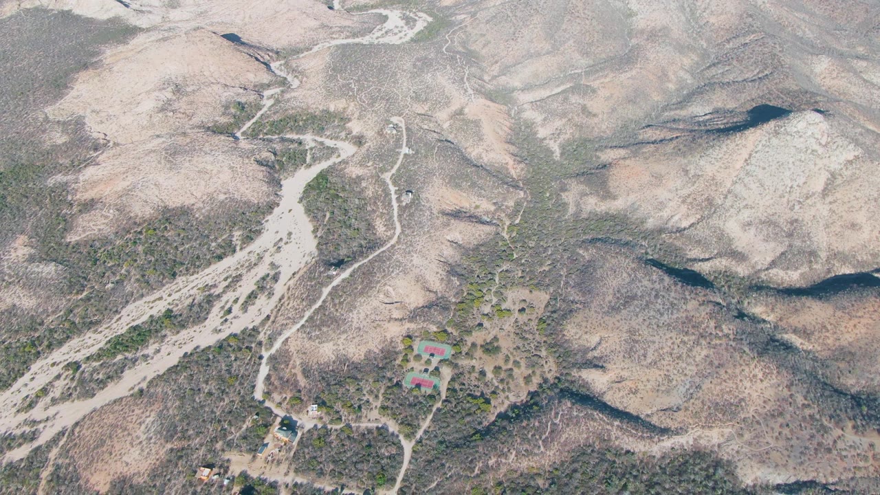 Aerial view in Cabo Pulmo shows the winding pale road bending through scrub-covered hills, with a small vehicle near the lower road area and no visible people.