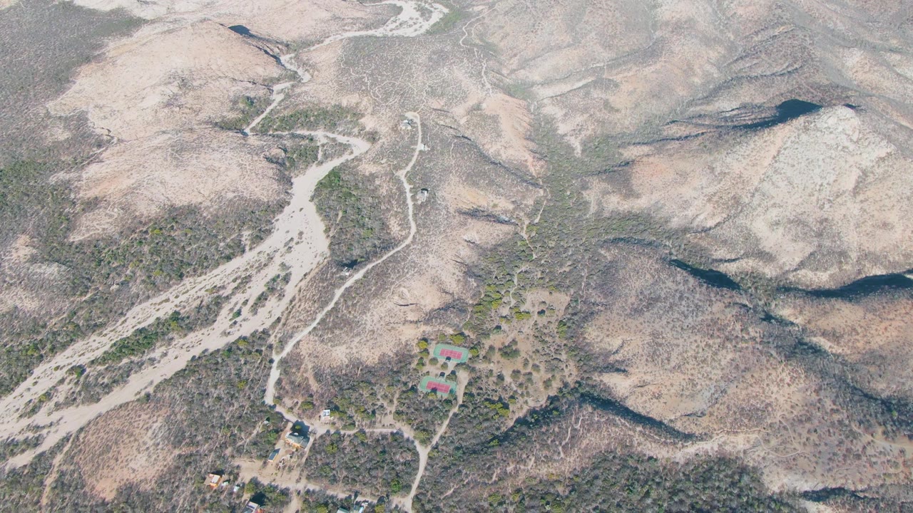 High drone-like frame over Cabo Pulmo shows pale dirt tracks threading between rocky slopes, scattered green scrub, small structures at the lower edge, and no visible people.