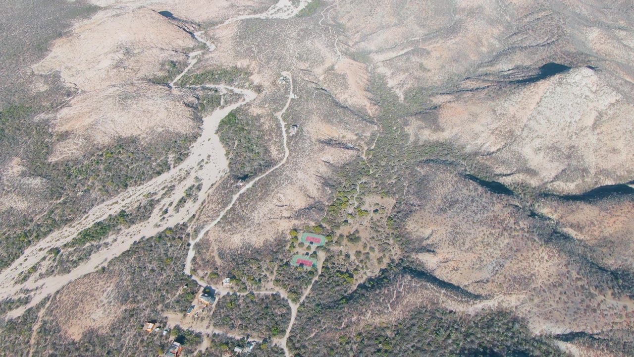 Overhead landscape view of a winding dirt road, desert wash, rocky hills, and a small property below.
