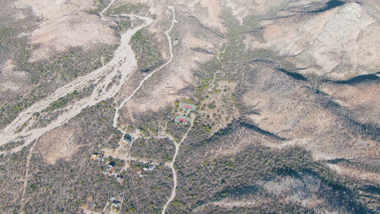 Wide aerial frame over Cabo Pulmo, Baja California Sur showing dry ridges, pale dirt tracks, small buildings, sparse shrubs, and no visible people.