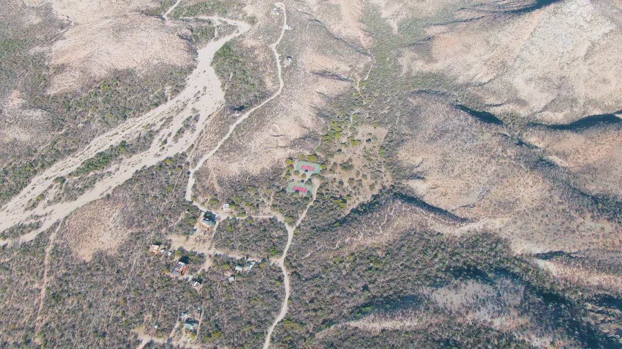 Aerial view in Cabo Pulmo, Baja California Sur with a pale wash crossing the upper left, a winding road near the center, hillside buildings, sparse vegetation, and no visible people.