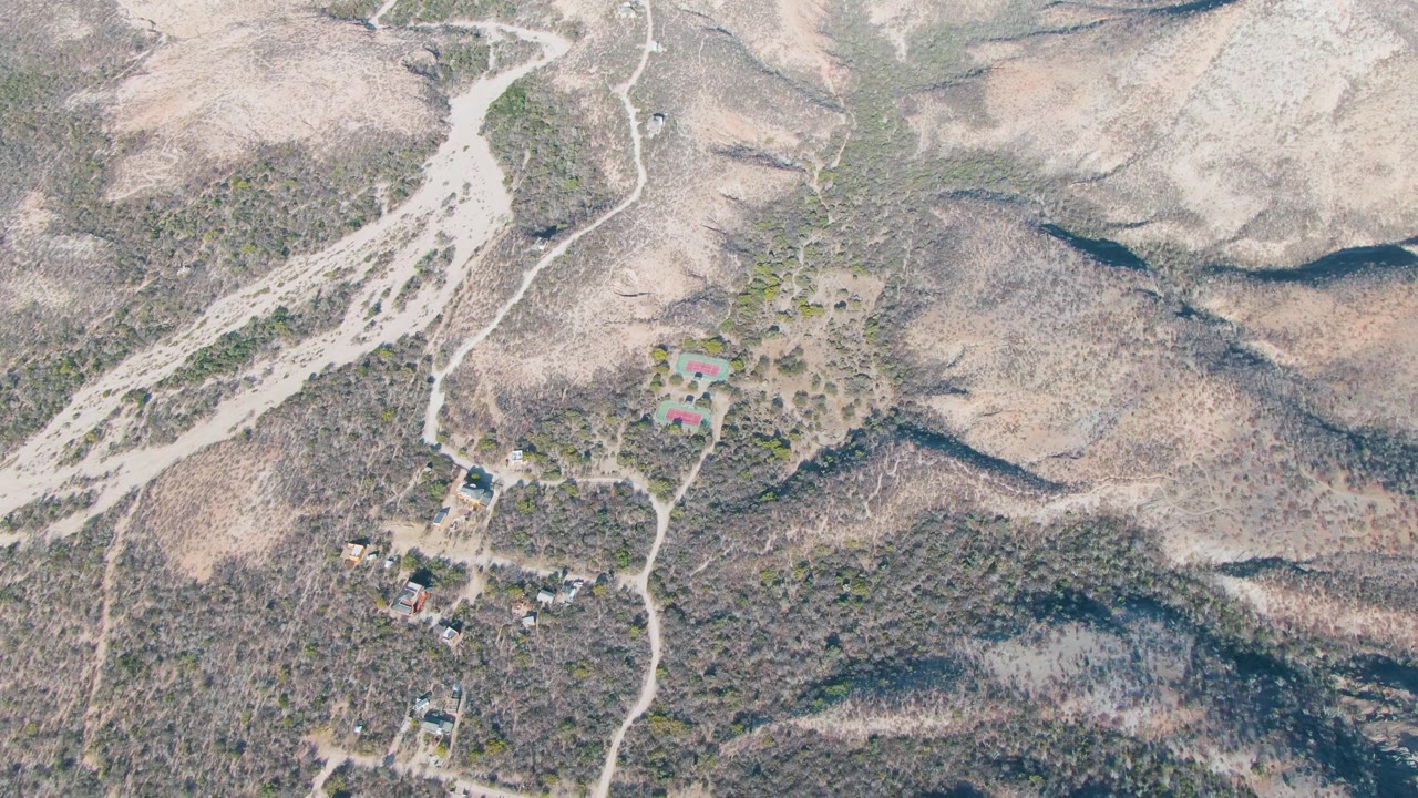 Drone-style overhead frame from Cabo Pulmo, Baja California Sur showing dusty roads splitting across dry hills, a small cluster of buildings, scattered green shrubs, and no visible people.