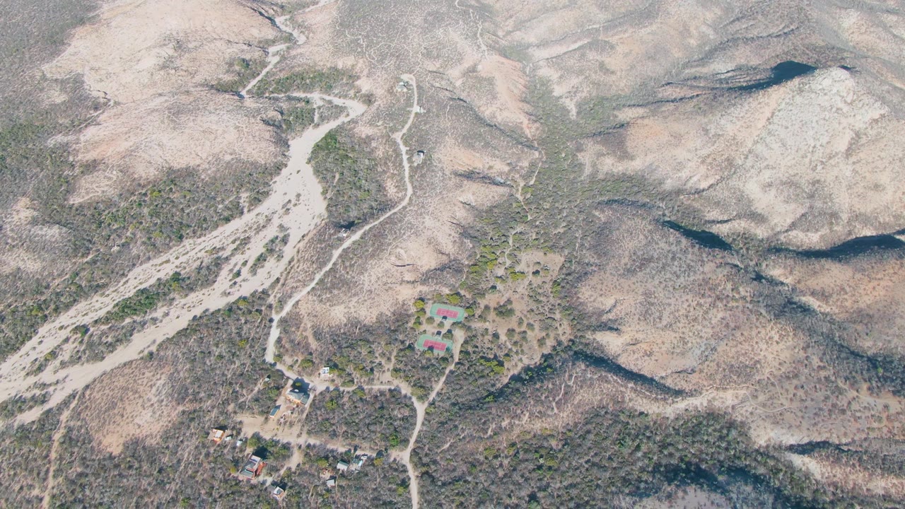 Opening aerial frame over Cabo Pulmo, Baja California Sur, showing pale dry hills, branching dirt roads, scattered rural buildings, sparse vegetation, and no visible people.