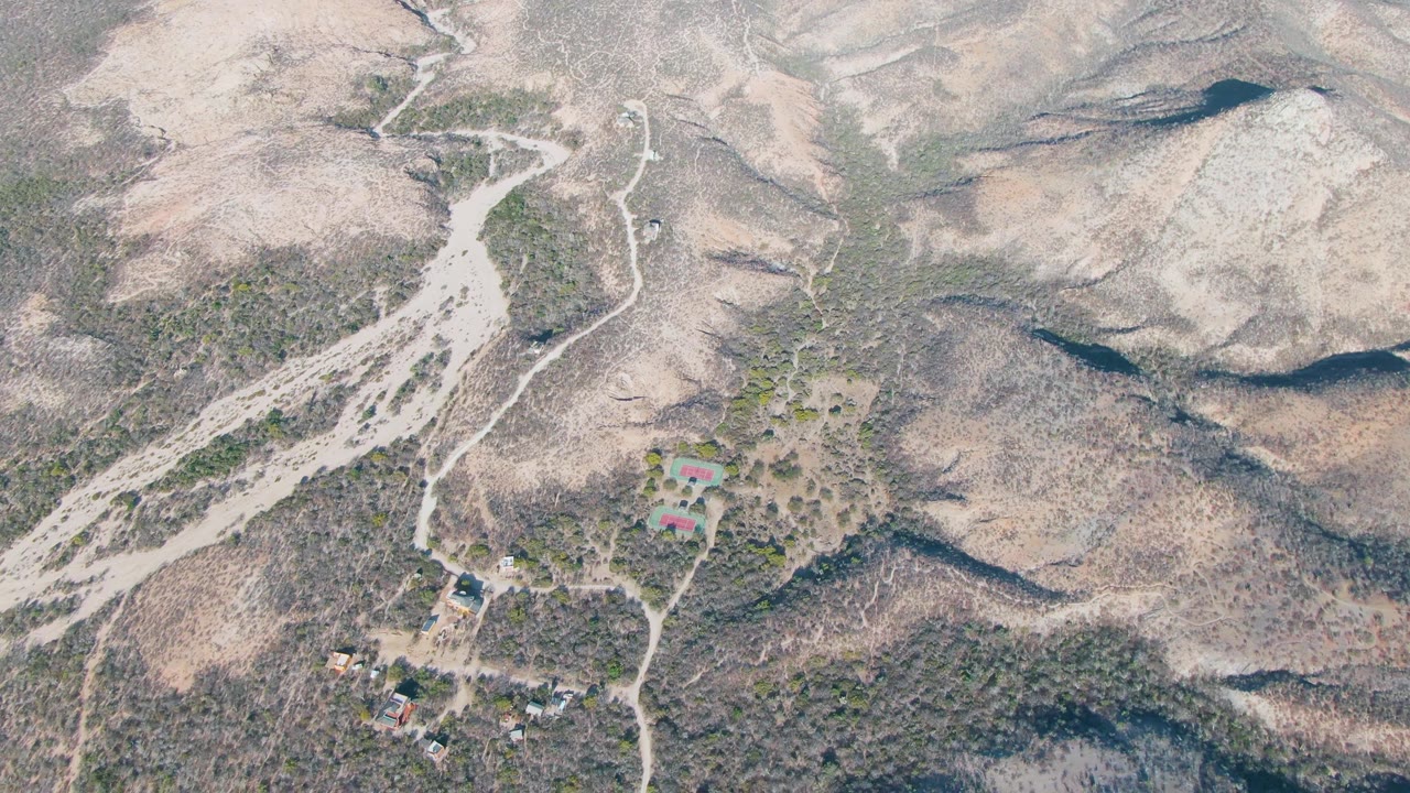 Drone view of winding dirt roads crossing rocky arid hills near scattered buildings.