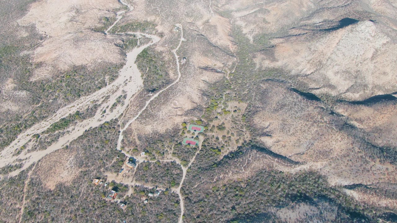 Mid-clip aerial frame over Cabo Pulmo, Baja California Sur, showing a dirt road corridor through dry hills, clustered rural buildings near the lower frame, and no visible people.