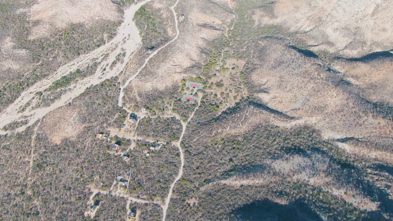 Aerial frame over Cabo Pulmo, Baja California Sur, showing dry hills, winding dirt tracks, scattered buildings, sparse brush, and no visible people at the start of the clip.
