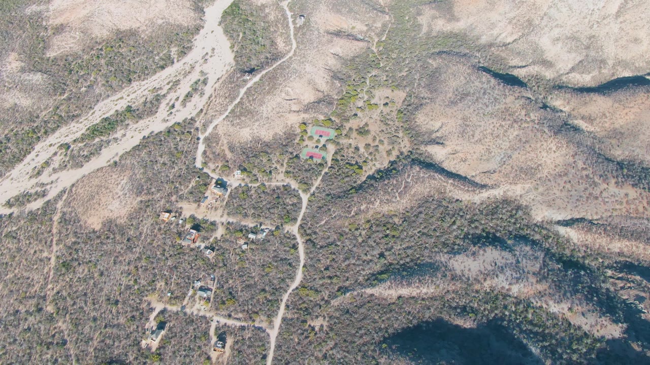 Aerial view over Cabo Pulmo, Baja California Sur, with a pale dirt road crossing dry slopes, small buildings set apart from each other, dark ridges, and no visible people.