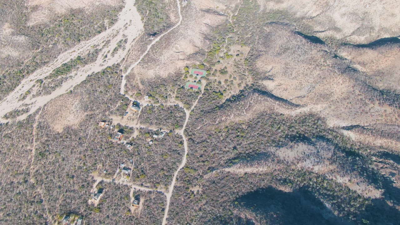 Drone frame over Cabo Pulmo, Baja California Sur, with diagonal ridgelines, branching dirt tracks, scattered rural buildings, scrub vegetation, and no visible people.