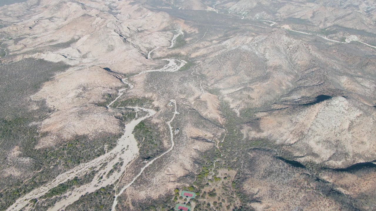 Opening aerial frame over Cabo Pulmo arid hills, with a pale dirt road curving through sparse scrub and no visible people.
