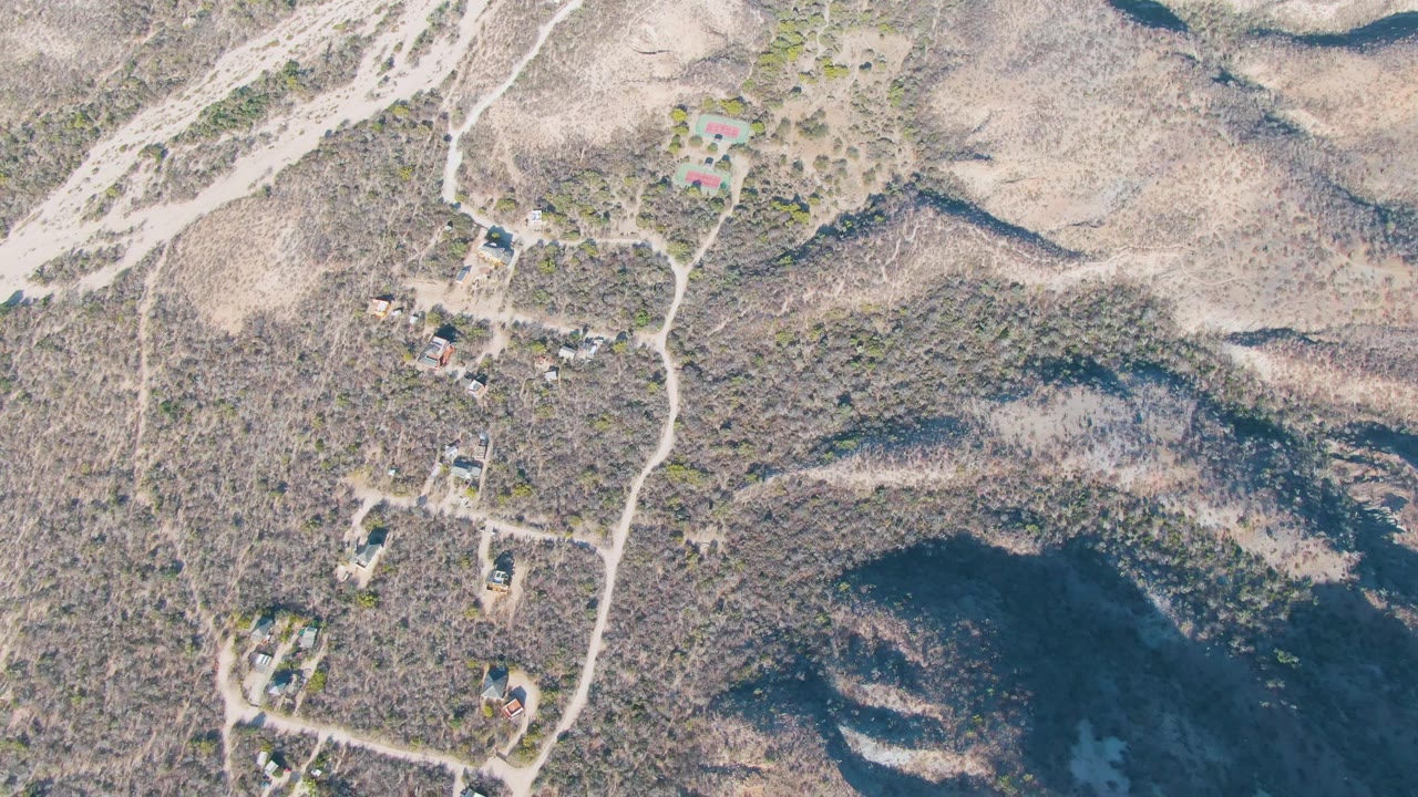 Overhead drone frame of arid hills near Cabo Pulmo, Baja California Sur, showing winding dirt roads, scattered buildings, sparse scrub, and no visible people.