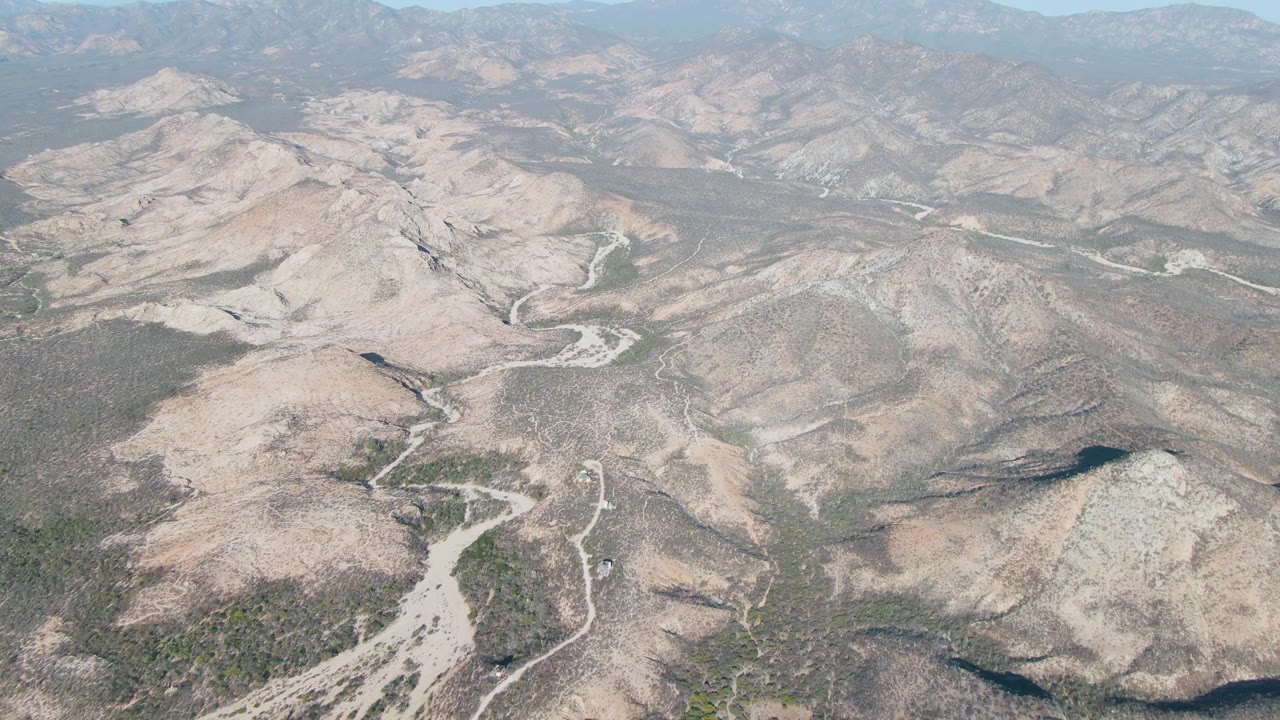 Wide aerial frame near Cabo Pulmo, Baja California Sur, with pale river washes crossing scrubby brown terrain and distant mountain ridges beneath a light blue haze, with no visible people.