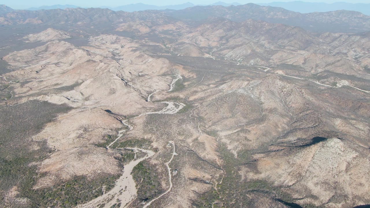 Aerial view near Cabo Pulmo, Baja California Sur, showing brighter rocky slopes around a winding dry wash, darker scrub patches, and layered hills in the distance, with no visible people.