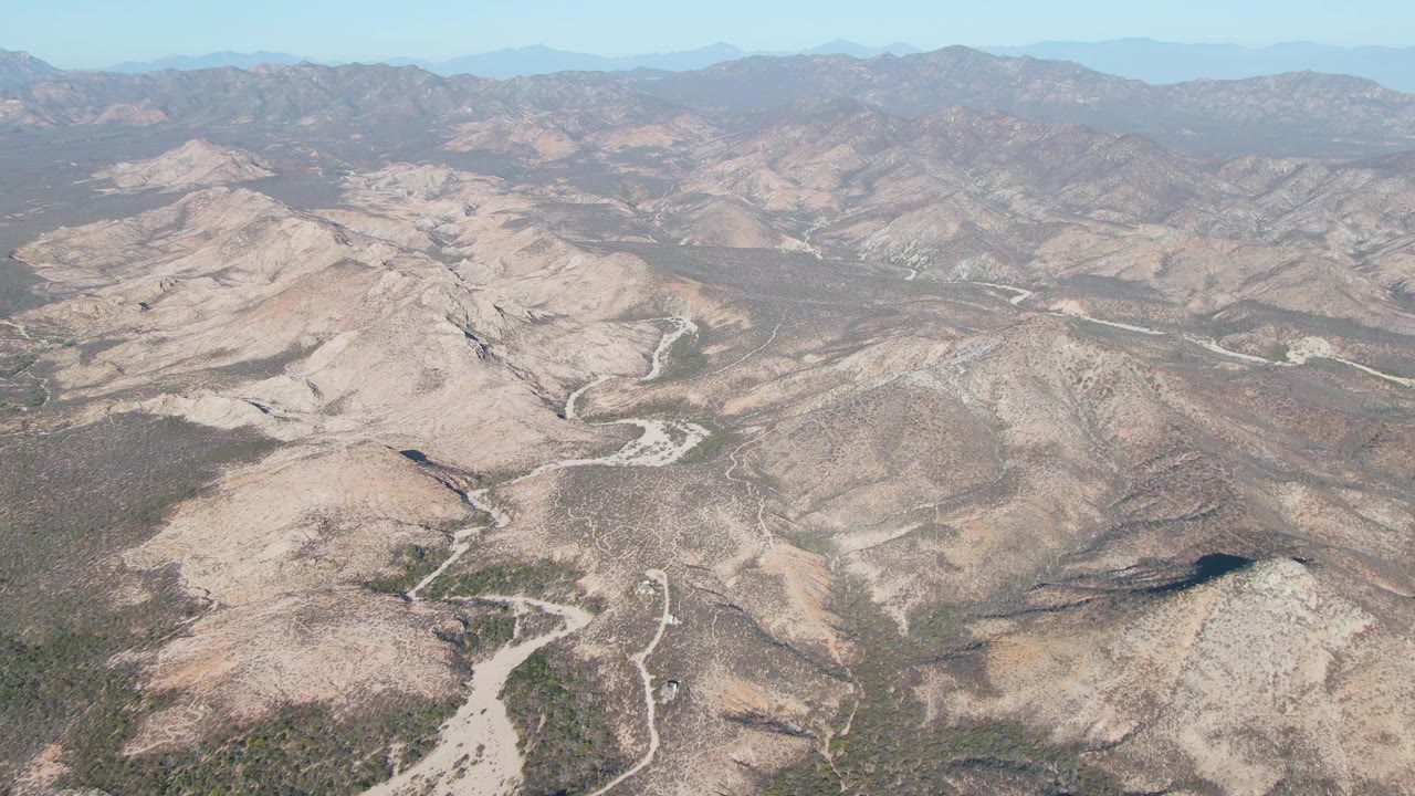 Aerial frame near Cabo Pulmo, Baja California Sur, looking across a dry valley of brown hills, pale river channels, sparse vegetation, and low mountain ranges, with no visible people.