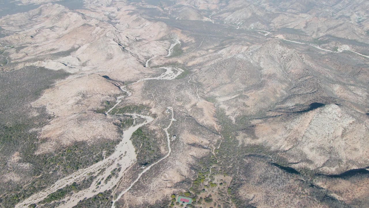 Elevated drone view in Cabo Pulmo showing a pale dry wash, curving dirt road, scrub-covered slopes, layered ridges, and no visible people.