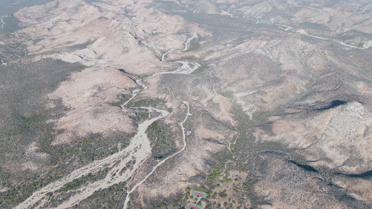 Drone view over Cabo Pulmo, Baja California Sur, with a branching pale riverbed cutting through dry hills and scrub patches, no visible people.