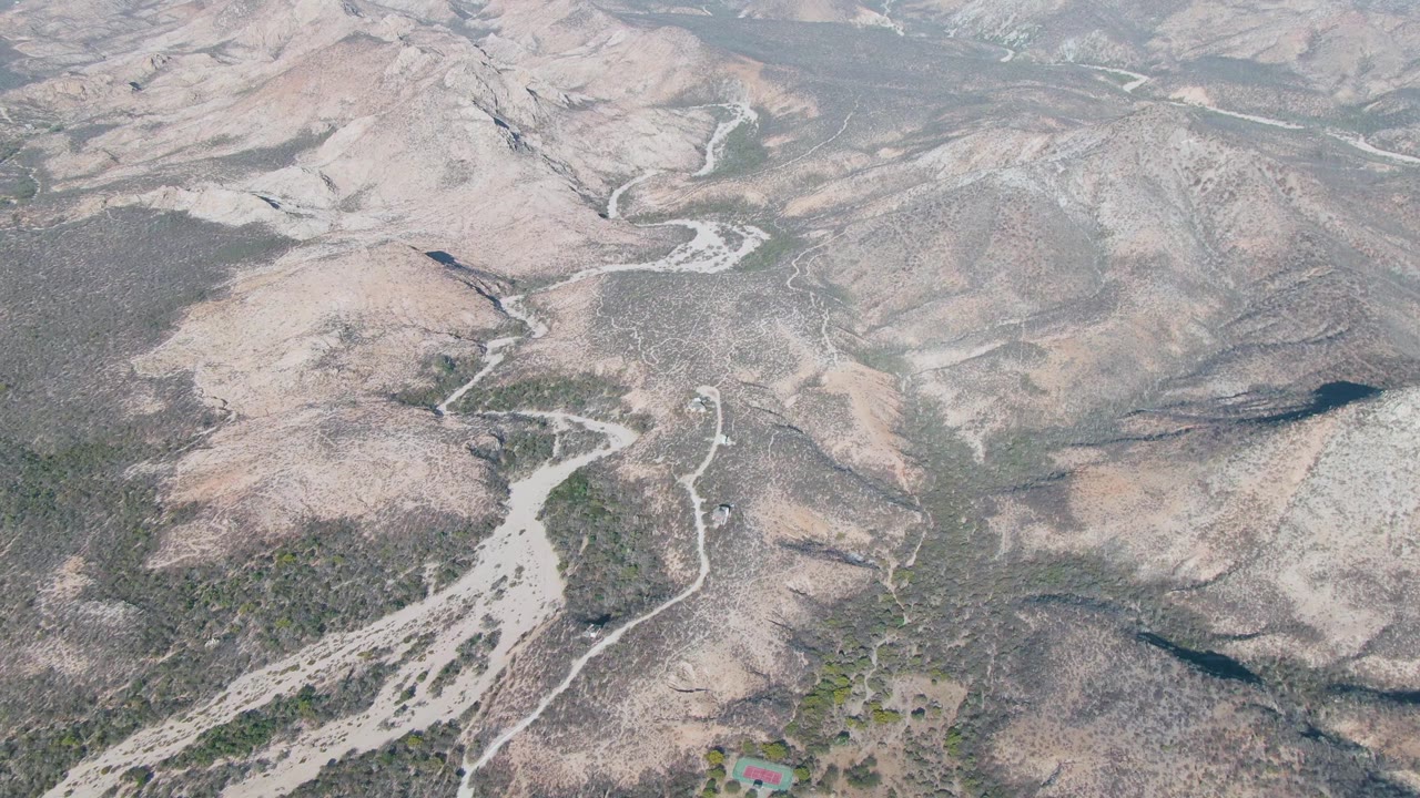 High aerial view in Cabo Pulmo, Baja California Sur, showing pale rocky slopes, a curving wash, and sparse green vegetation across dry mountain terrain with no visible people.