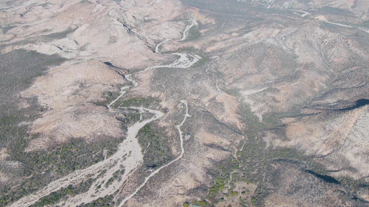 Aerial view over Cabo Pulmo, Baja California Sur, with the pale wash nearer the lower left, scrub-covered terrain, and rounded dry hills beyond, no visible people.