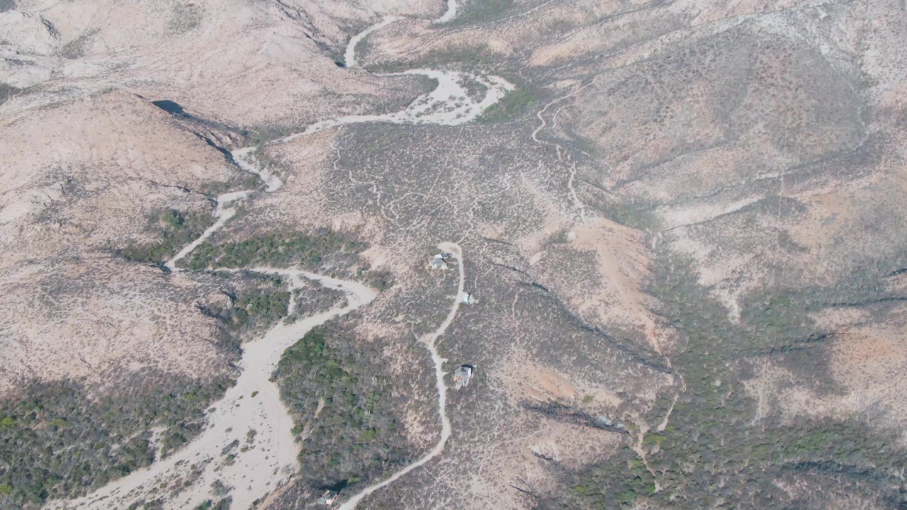 Drone-like overhead frame from Cabo Pulmo, Baja California Sur showing a winding pale channel beside darker scrub patches on dry hilly ground with no visible people.