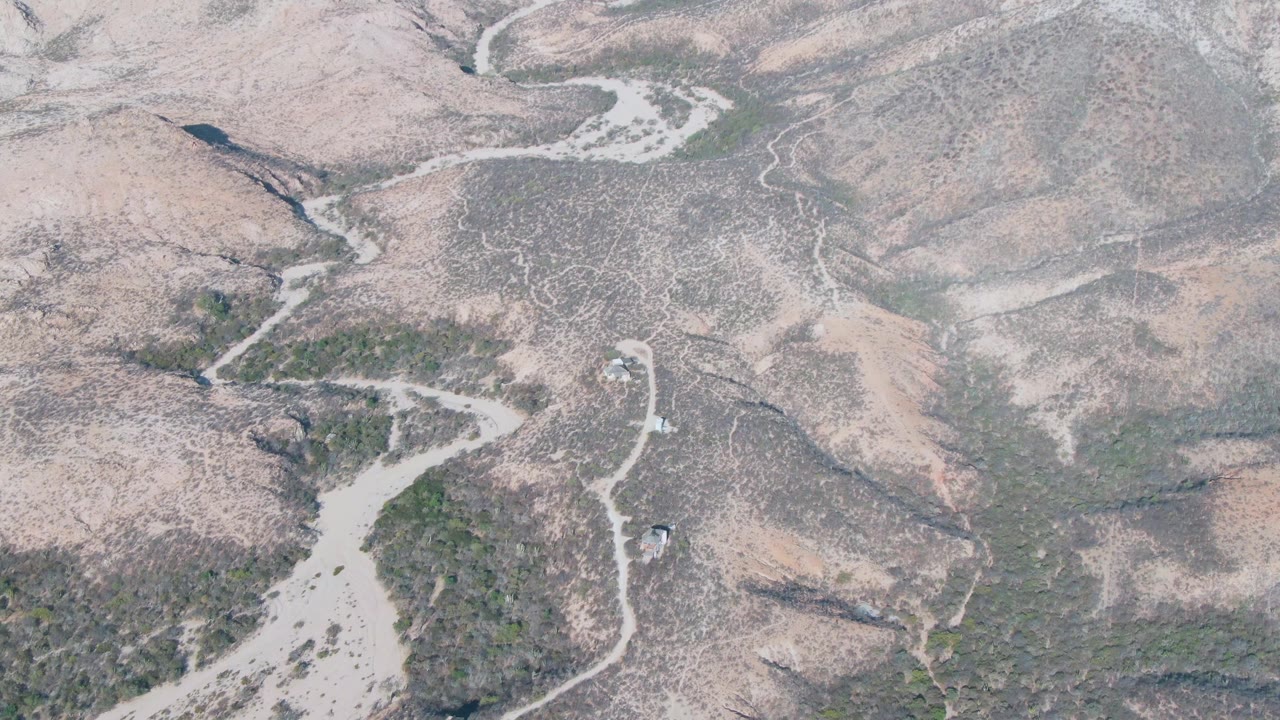 Aerial frame over the Cabo Pulmo area showing a light riverbed through a dry valley, green patches along the drainage, rocky hillsides, and no visible people.