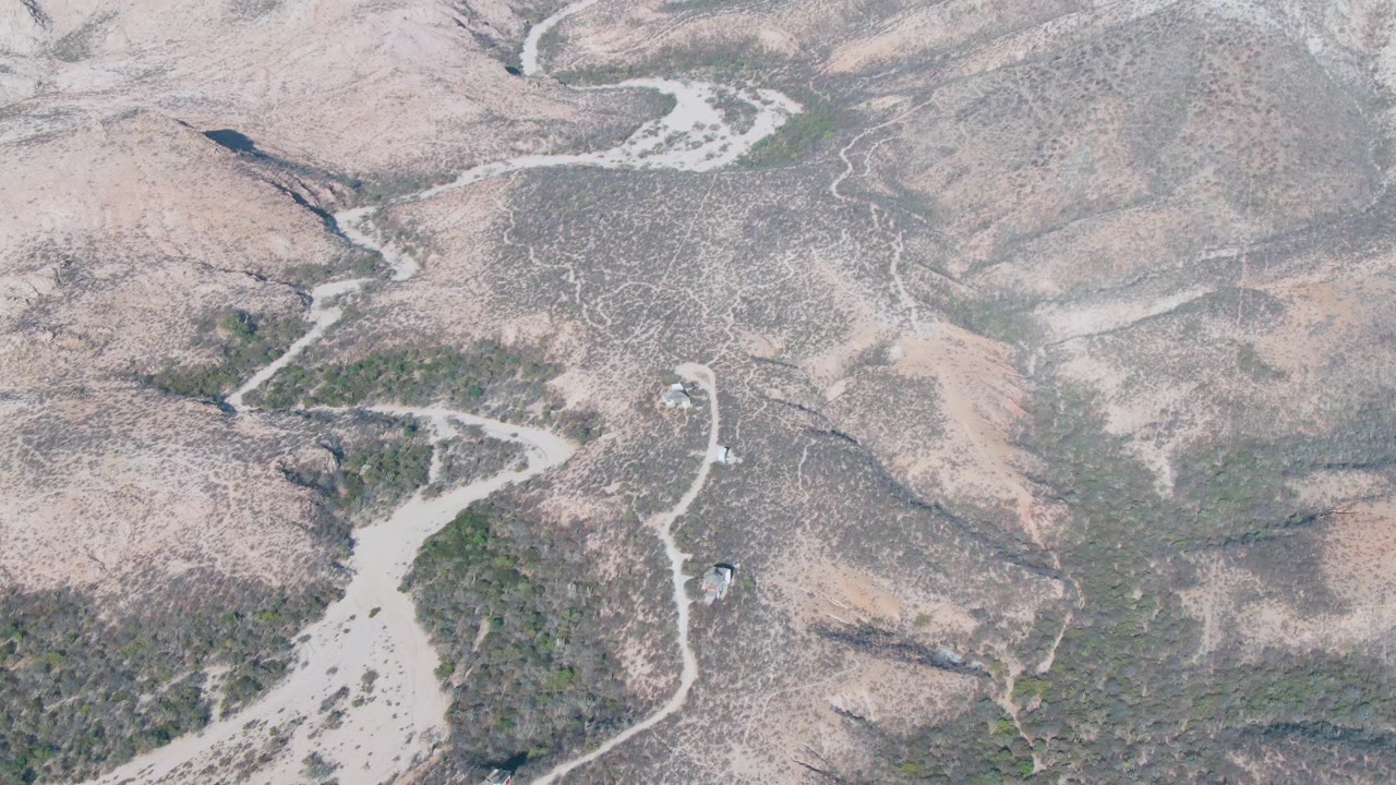 Aerial view over Cabo Pulmo arid hills with the vehicle farther along the pale road, dry washes cutting across the terrain, and no visible people.