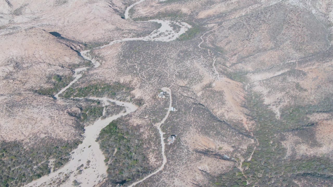 Drone-style overhead frame in Cabo Pulmo with a small vehicle near a winding road, muted tan and gray slopes, patches of green vegetation, and no visible people.