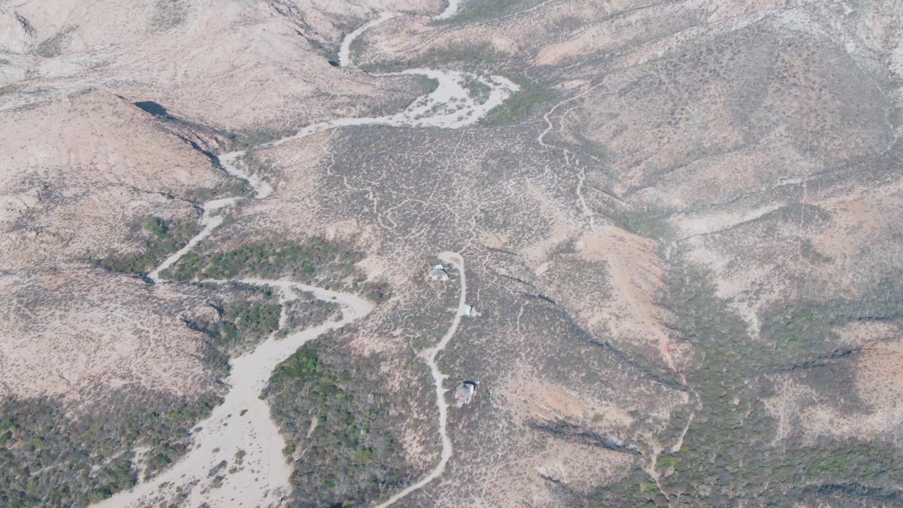 Aerial frame from Cabo Pulmo showing the vehicle beside a pale road segment, with a sandy wash at lower left, dry ridges, sparse shrubs, and no visible people.