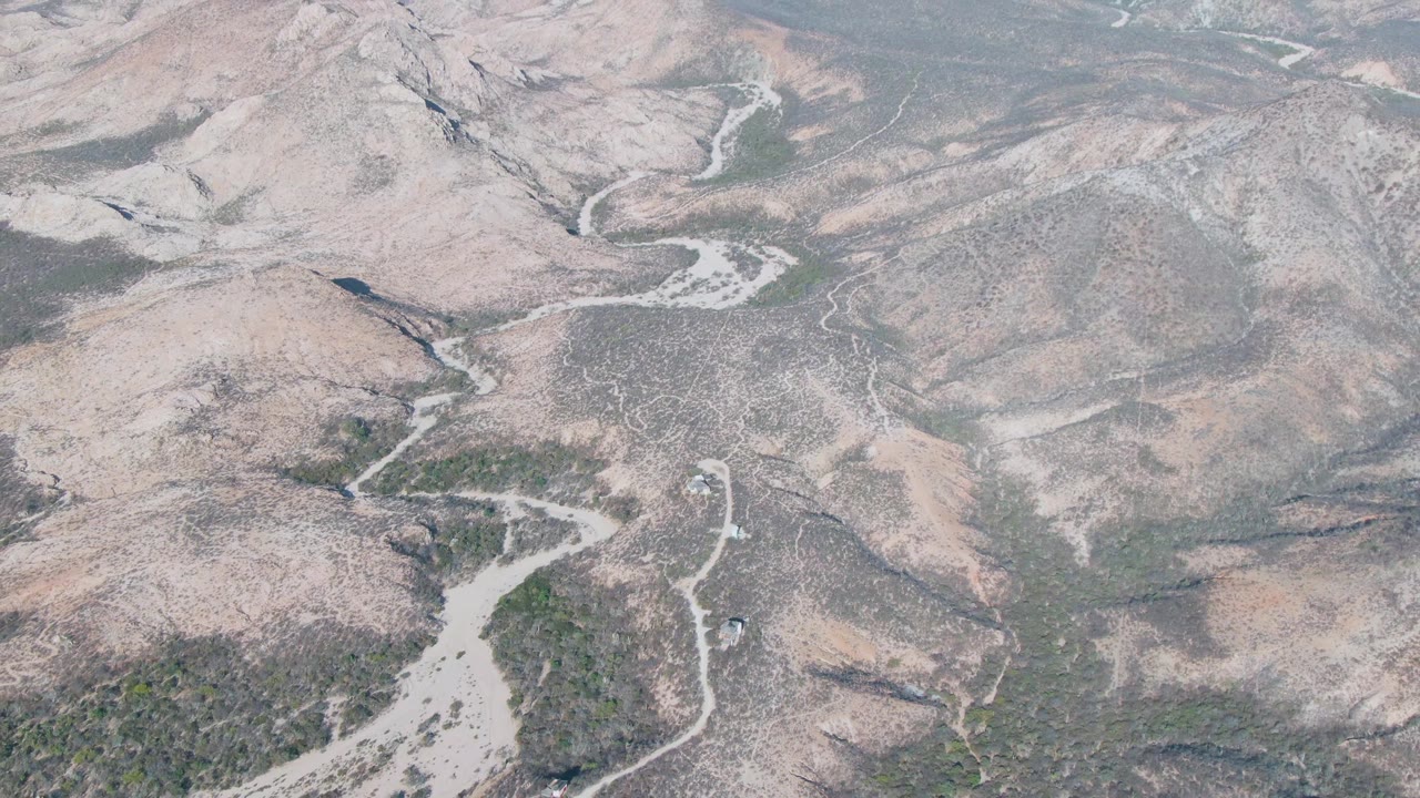 Opening aerial frame over Cabo Pulmo arid hills with pale dry riverbeds, rocky slopes, sparse green vegetation, and no visible people.