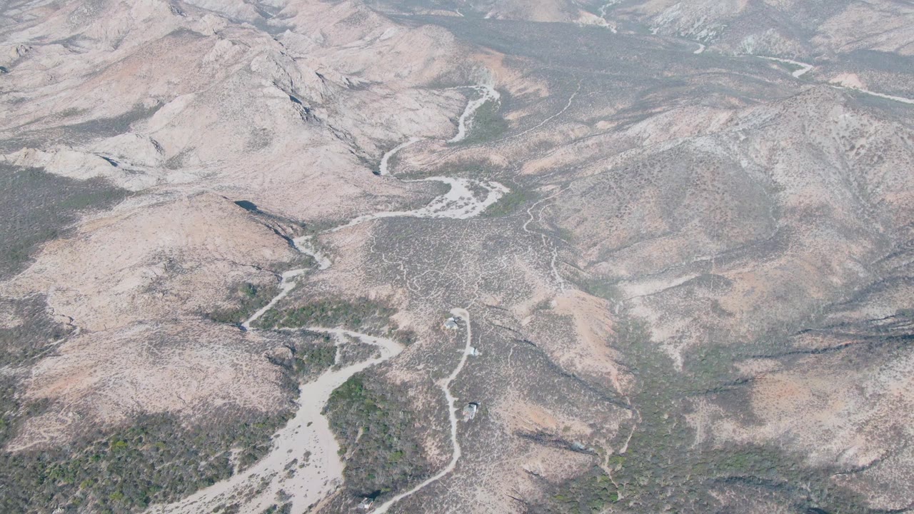 Aerial frame over Cabo Pulmo showing a winding pale wash between brown and tan hills, scattered green scrub, hazy distance, and no visible people.