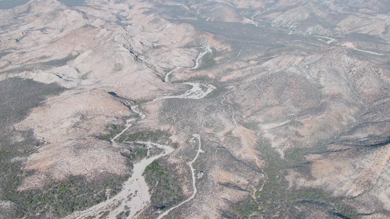 Mid-clip aerial over Cabo Pulmo terrain with branching dry riverbeds, pale gravel channels, muted brown hills, sparse vegetation patches, and no visible people.