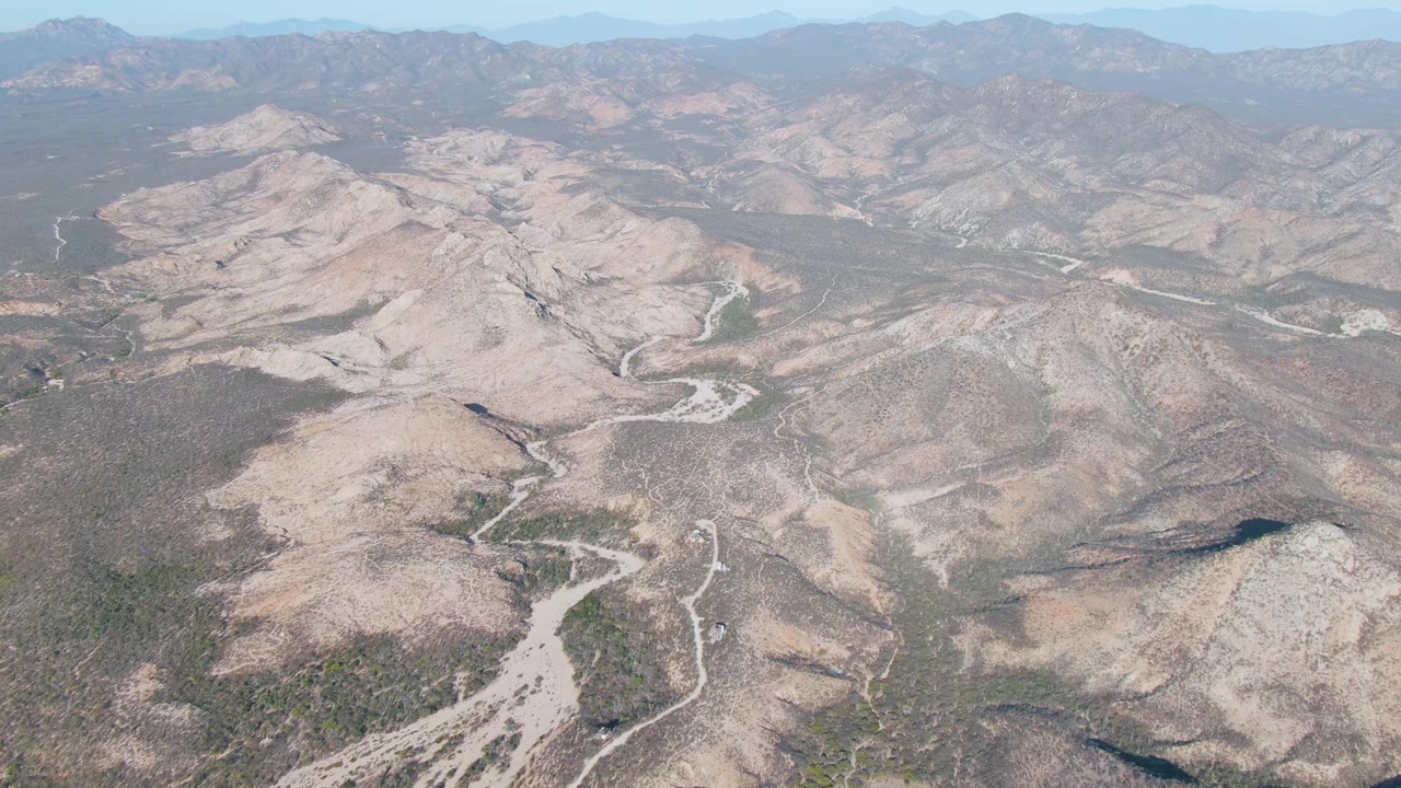 Opening aerial frame over Cabo Pulmo East Cape desert hills with pale dry washes, sparse vegetation, distant hazy mountains, and no visible people.