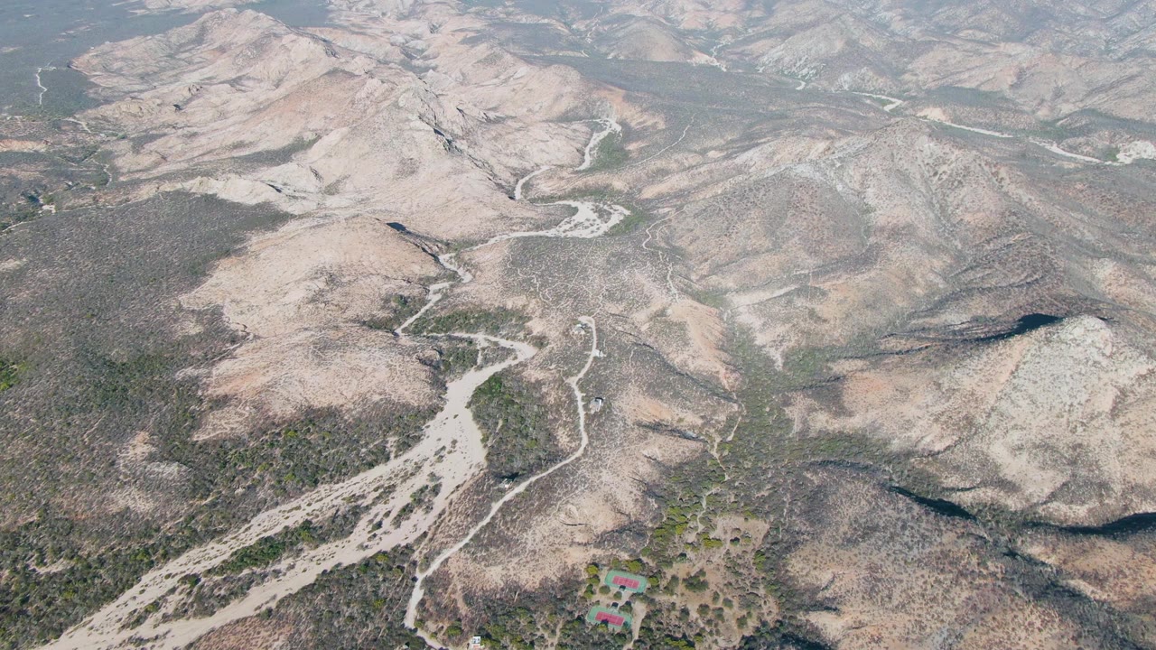A later aerial view of Cabo Pulmo hillside terrain shows the dry wash branching across brown slopes, with scrub vegetation, faint dirt routes, a small red-roofed structure near the bottom edge, and no visible people.