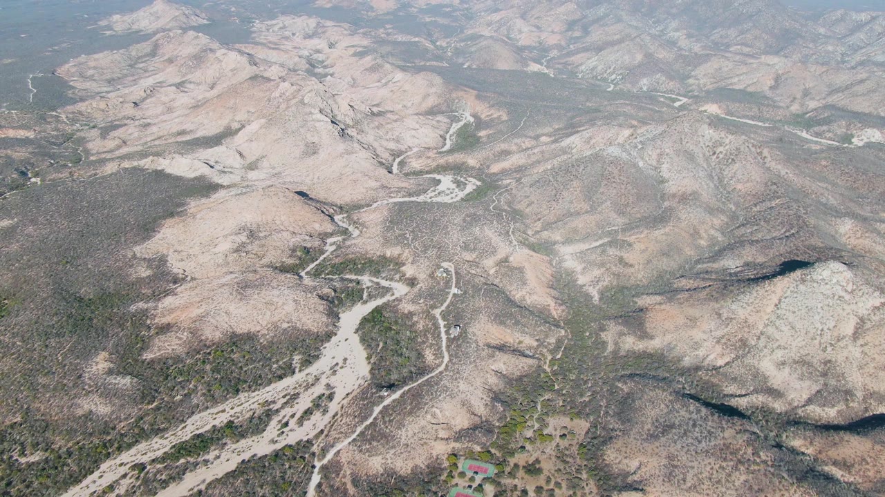 Aerial view over Cabo Pulmo dry mountain terrain with pale rocky ridges, sparse vegetation, a winding dirt road in the valleys, hazy distant hills, and no visible people.